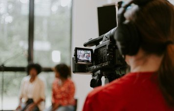 A foto é tirada por trás de uma operadora de câmera (vestindo uma camiseta vermelha e fones de ouvido), colocando o espectador no papel de quem produz o conteúdo.