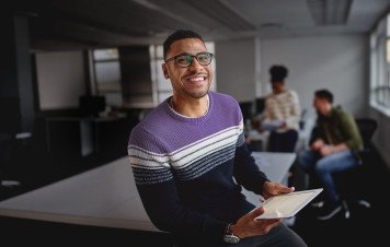 Um homem jovem, negro, com óculos e um sorriso expressivo, posa para a câmera. Ele veste um suéter listrado em tons de roxo, branco e azul marinho e segura um tablet, sugerindo o uso de tecnologia no dia a dia.