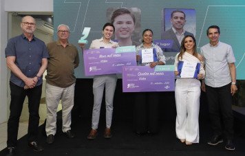 Um grupo de seis pessoas posa para a foto em um palco. No centro, os premiados seguram cheques simbólicos de grande formato (um no valor de 9 mil reais e outro de 4 mil reais), certificados e um troféu em formato de aspas.