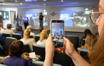 Uma pessoa segura um smartphone em destaque, gravando ou transmitindo ao vivo o que acontece no palco. A tela do celular mostra exatamente a cena à frente. Em encontro com a imprensa, Sebrae/SC apresenta a 13ª edição do Prêmio Sebrae de Jornalismo