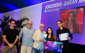 Um grupo de cinco pessoas está posicionado no palco. No centro, uma jovem sorridente recebe um troféu em formato de aspas e segura um cheque simbólico no valor de mil e quinhentos reais. Ao seu lado direito, um jovem segura o certificado de premiação.