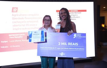 Duas mulheres sorridentes posam no palco segurando os símbolos da vitória. A mulher à direita, com cabelos trançados, segura um troféu em formato de aspas e um cheque simbólico no valor de 2 mil reais pelo 2º lugar no prêmio. A mulher à esquerda, de óculos, segura o certificado oficial.