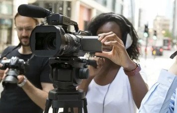 Uma mulher negra, utilizando fones de ouvido profissionais, opera uma câmera de vídeo de grande porte montada em um tripé. Ela ajusta o visor lateral do equipamento com atenção.