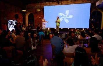 Esta imagem registra uma cerimônia ou apresentação oficial realizada em um espaço de eventos com estética rústica e elegante. Um homem de camisa verde fala ao microfone no centro do palco, enquanto uma mulher de vestido claro está ao lado em um púlpito acrílico transparente.