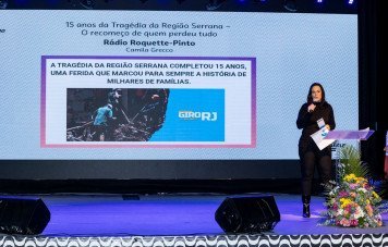 Esta imagem registra a apresentação de um trabalho jornalístico durante uma cerimônia ou evento oficial. À direita, uma mulher vestindo roupa preta fala ao microfone em um púlpito acrílico decorado com flores coloridas. Um grande painel exibe a apresentação da reportagem com o título: 