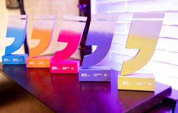 Esta imagem exibe cinco troféus da 12ª Edição do Prêmio Sebrae de Jornalismo, alinhados sobre uma bancada de madeira escura. Cada troféu possui o formato característico de aspas estilizadas com gradiente de cor no topo e base retangular gravada com as identificações da premiação.