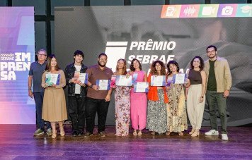 Esta imagem é uma fotografia oficial registrada durante a cerimônia de entrega do Prêmio Sebrae de Jornalismo. Dez pessoas (vencedores e representantes) estão alinhadas no centro do palco, posando sorridentes para a foto. Os premiados exibem certificados de participação e troféus em formato de aspas estilizadas com bases coloridas. Ao fundo, um grande telão exibe o logotipo da premiação acompanhado pelos ícones coloridos das modalidades no canto superior direito. No painel à esquerda, lê-se a marca 