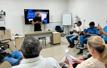 Esta imagem registra uma reunião presencial ou sessão informativa sobre o Prêmio Sebrae de Jornalismo, realizada em uma sala de reuniões. Ao fundo, uma grande TV na parede exibe o slide oficial da campanha do prêmio, com o slogan 