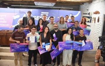 Um grupo de premiados do **Prêmio Sebrae de Jornalismo** posa para foto reunido em um evento. Os participantes sorriem enquanto exibem troféus, certificados e cheques simbólicos de premiação em dinheiro em frente a um painel institucional do Sebrae.