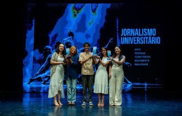 Cinco premiados posam juntos no centro do palco durante uma cerimônia de premiação de jornalismo. Eles sorriem e exibem troféus e certificados em frente a um grande painel digital azul que destaca a inscrição **