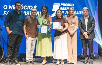 Esta imagem registra a entrega de premiação durante a cerimônia do Prêmio Sebrae de Jornalismo. Seis pessoas posam lado a lado em tom de celebração no centro do palco. Ao centro, duas mulheres seguram com orgulho seus certificados oficiais e um troféu em acrílico. A mulher à esquerda veste um vestido verde-claro e a da direita usa saia branca com blusa marrom.