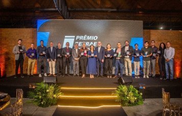 Foto de grupo com dezoito pessoas alinhadas lado a lado em um palco decorado, durante a cerimônia do Prêmio Sebrae de Jornalismo. Quase todos os presentes vestem trajes esporte fino ou formais e seguram troféus de premiação nas mãos. Ao fundo, destaca-se um painel azul e preto com a inscrição iluminada 