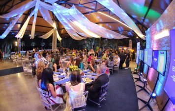 A imagem mostra a visão ampla do interior de um amplo salão de festas decorado para um evento formal à noite. O teto alto possui estrutura metálica coberta por tecidos brancos drapeados, com iluminação cênica em tons azuis e coloridos. Ao lado direito, há uma parede de tijolos aparentes com monitores digitais instalados em suportes.