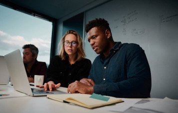 Uma equipe trabalhando em um escritório moderno, com destaque para dois colegas concentrados na tela de um notebook. Ao fundo, observam-se anotações em um quadro-branco e outro profissional em seu posto de trabalho.