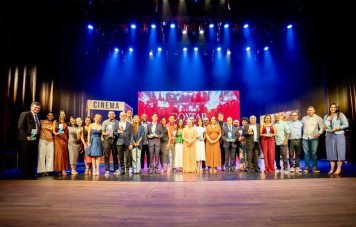 Um grande grupo de homenageados e organizadores posa alinhado no palco de um teatro durante uma cerimônia de premiação. As pessoas vestem trajes esporte fino e de gala, com várias delas segurando troféus e certificados, sob iluminação cênica azul e um painel ao fundo com a frase 