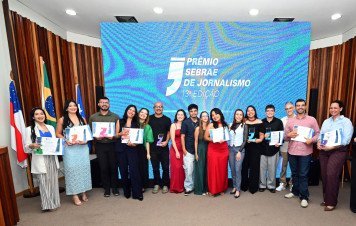 Esta imagem registra a fotografia oficial coletiva dos premiados e finalistas durante a etapa estadual do Prêmio Sebrae de Jornalismo (13ª Edição). Cerca de 17 pessoas posam alinhadas lado a lado em tom de comemoração no centro do palco. Os participantes exibem com orgulho seus certificados de premiação e troféus em acrílico em formato de aspas com bases coloridas.
