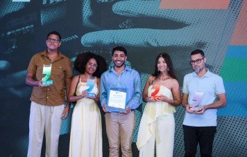 Esta imagem registra a premiação dos vencedores das diversas categorias durante uma cerimônia do Prêmios Sebrae de Jornalismo. Cinco jornalistas posam alinhados no centro do palco, exibindo sorridentes seus reconhecimentos.
