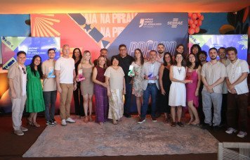 Esta imagem registra a foto coletiva dos premiados, finalistas e organizadores durante a cerimônia da 13ª Edição do Prêmio Sebrae de Jornalismo - Etapa Distrito Federal. Cerca de 20 pessoas (jornalistas finalistas, vencedores e representantes) posam juntas e sorridentes no espaço do evento. Diversos premiados exibem seus troféus em acrílico com o formato característico de aspas e bases coloridas representativas das modalidades da premiação.