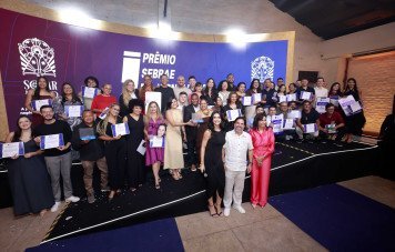 Esta imagem registra a fotografia coletiva oficial dos premiados, finalistas e organizadores durante uma cerimônia do Prêmio Sebrae de Jornalismo. Um grande grupo composto por mais de 30 pessoas posa sorridente no palco e à frente dele. Os jornalistas e homenageados exibem com orgulho seus certificados de premiação emoldurados e troféus em acrílico.