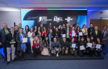 Esta imagem registra a fotografia oficial coletiva dos vencedores, finalistas e comissão organizadora da Etapa Paraná da 13ª Edição do Prêmio Sebrae de Jornalismo. Um grande grupo com cerca de 30 pessoas posa sorridente, organizado em duas fileiras (em pé e agachadas na frente) Os participantes exibem com orgulho seus troféus em acrílico e certificados oficiais de premiação. Ao fundo, um telão de LED institucional destaca a marca do Prêmio Sebrae de Jornalismo - 13ª Edição - Etapa Paraná, acompanhado dos logotipos da ASN (Agência Sebrae de Notícias) e do Sebrae.