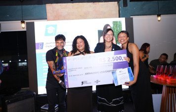 Esta imagem registra o momento da premiação da categoria Jornalismo Universitário durante uma cerimônia do Prêmio Sebrae de Jornalismo. Quatro pessoas sorridentes posam juntas no centro do palco segurando um cheque simbólico gigante no valor de R$ 2.500,00, nominal à categoria de Jornalismo Universitário.
