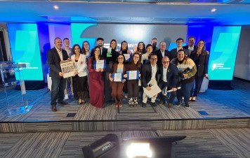 Esta imagem registra a foto coletiva dos premiados e organizadores durante a etapa estadual (Santa Catarina) do Prêmio Sebrae de Jornalismo. Cerca de 20 pessoas (vencedores, finalistas e representantes) posam juntas no palco em um momento de comemoração. Os homenageados exibem seus certificados oficiais e troféus em acrílico. No canto esquerdo, um participante segura uma edição impressa promocional tipo jornal (