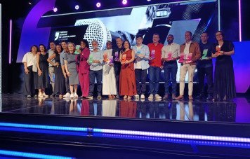 Esta imagem registra a fotografia oficial coletiva dos premiados e homenageados durante a cerimônia da 13ª Edição do Prêmio Sebrae de Jornalismo. Cerca de 16 profissionais e convidados posam alinhados lado a lado ao longo de um amplo palco brilhante e espelhado. Os vencedores exibem troféus em acrílico com bases coloridas vibrantes (verde, laranja, rosa e roxo), representativas das diversas categorias da premiação.