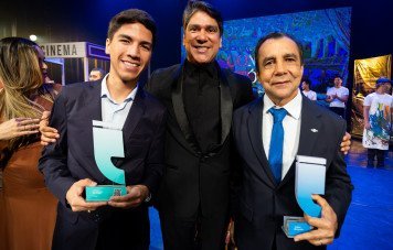 Esta imagem registra o momento de celebração e entrega de prêmios em um palco durante a cerimônia do Prêmio Sebrae de Jornalismo. Três homens posam sorridentes e abraçados no primeiro plano. Ao centro, um apresentador/organizador vestindo blazer preto; à esquerda, um jovem premiado vestindo terno escuro e camisa clara; e à direita, um homem de terno com gravata azul. O premiado à esquerda segura o troféu em acrílico em formato de aspas com base em tom ciano/verde (categoria de vídeo), enquanto o homenageado à direita exibe um troféu semelhante com base azul.