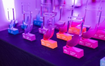 Esta imagem exibe em detalhe um conjunto de troféus oficiais do Prêmio Sebrae de Jornalismo, organizados sobre uma mesa antes ou durante a cerimônia de entrega. As peças em acrílico transparente possuem o formato icônico de aspas estilizadas, assentadas sobre bases retangulares coloridas que identificam a colocação e a categoria (como 