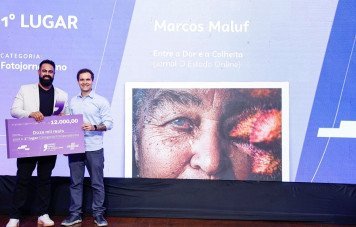 Esta imagem registra o momento da premiação do 1º Lugar na Categoria Fotojornalismo durante uma cerimônia do Prêmio Sebrae de Jornalismo. Participantes no Palco: Dois homens posam sorridentes no centro do palco, celebrando a conquista do prêmio. Reconhecimentos e Cheque: O premiado à esquerda segura um cheque simbólico gigante no valor de R$ 12.000,00 (doze mil reais) referente ao primeiro lugar na categoria Telão e Informações do Trabalho: Ao fundo, o painel exibe a fotografia vencedora, indicando o nome do autor Marcos Maluf e o título do trabalho 