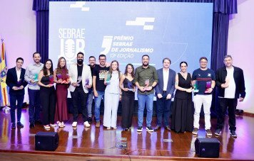 Esta imagem registra a fotografia oficial coletiva dos premiados e organizadores durante a etapa Mato Grosso do Sul da 13ª Edição do Prêmio Sebrae de Jornalismo. Cerca de 15 profissionais posam alinhados no centro do palco em clima festivo e de celebração. Os premiados exibem troféus em acrílico com o formato de aspas e bases coloridas (verde, rosa, roxo e azul), representativas das modalidades da edição.
