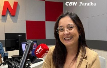 Esta imagem registra o retrato individual de uma jornalista nos estúdios da rádio CBN Paraíba. Uma mulher sorridente com óculos de armação fina, blazer bege e blusa preta posa sentada à bancada de transmissão. Em primeiro plano, destaca-se o microfone profissional com a espuma vermelha personalizada com a marca CBN Paraíba, além de mesa de som e telefones de estúdio ao fundo. Na parede ao fundo, observa-se a cenografia da emissora com painéis quadriculados em vermelho e cinza e a inscrição 