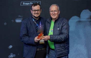 Esta imagem registra o momento da entrega de prêmio durante a etapa Paraná do Prêmio Sebrae de Jornalismo. Dois homens posam sorridentes lado a lado ao centro do palco. À esquerda, o jornalista premiado vestindo blazer azul-escuro e óculos; à direita, o representante da organização vestindo jaqueta escura sobre camiseta verde com a marca do Sebrae. Ambos seguram juntos o troféu oficial em acrílico transparente em formato de aspas com base em tom laranja/avermelhado. Ao fundo, um painel escuro exibe a identificação da etapa com o texto parcial 