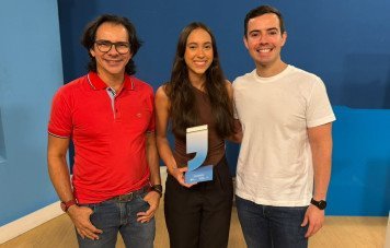 Esta imagem registra a comemoração de uma conquista com a entrega de troféu no contexto do Prêmio Sebrae de Jornalismo - Etapa Rio Grande do Norte. Participantes: Três pessoas posam sorridentes lado a lado em um estúdio. À esquerda, um homem com óculos e camisa polo vermelha; ao centro, uma jovem segurando o troféu; à direita, um homem vestindo camiseta branca e calça jeans. Troféu em Destaque: A participante ao centro segura o troféu oficial em acrílico transparente em formato de aspas com base em tom azul e identificação de vencedora. Cenário e Ambiência: O ambiente interno possui paredes em tons de azul e piso amadeirado, transmitindo um clima descontraído e de orgulho pela premiação.