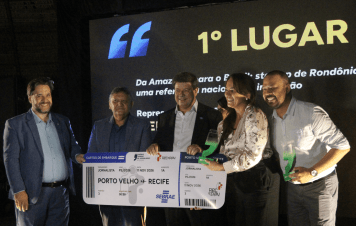 Esta imagem registra a entrega da premiação do 1º Lugar durante a cerimônia da etapa Rondônia do Prêmio Sebrae de Jornalismo. Cinco pessoas posam sorridentes no centro do palco durante a entrega do prêmio principal. Os vencedores exibem troféus em acrílico com base verde e seguram um cartão de embarque simbólico gigante (trecho Porto Velho - Recife) representando a passagem para a próxima etapa. A fotografia captura um momento de celebração, conquista e reconhecimento do jornalismo regional.
