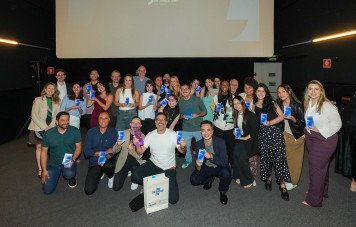 Esta imagem registra a fotografia oficial coletiva dos premiados e finalistas durante a etapa São Paulo do Prêmio Sebrae de Jornalismo. Um grande grupo com cerca de 25 pessoas posa sorridente no auditório, distribuído em pé e ajoelhado à frente. Diversos participantes exibem troféus em acrílico com a base azul e certificados/placas institucionais. À frente, no chão, destaca-se também uma sacola promocional com o logotipo do Sebrae.